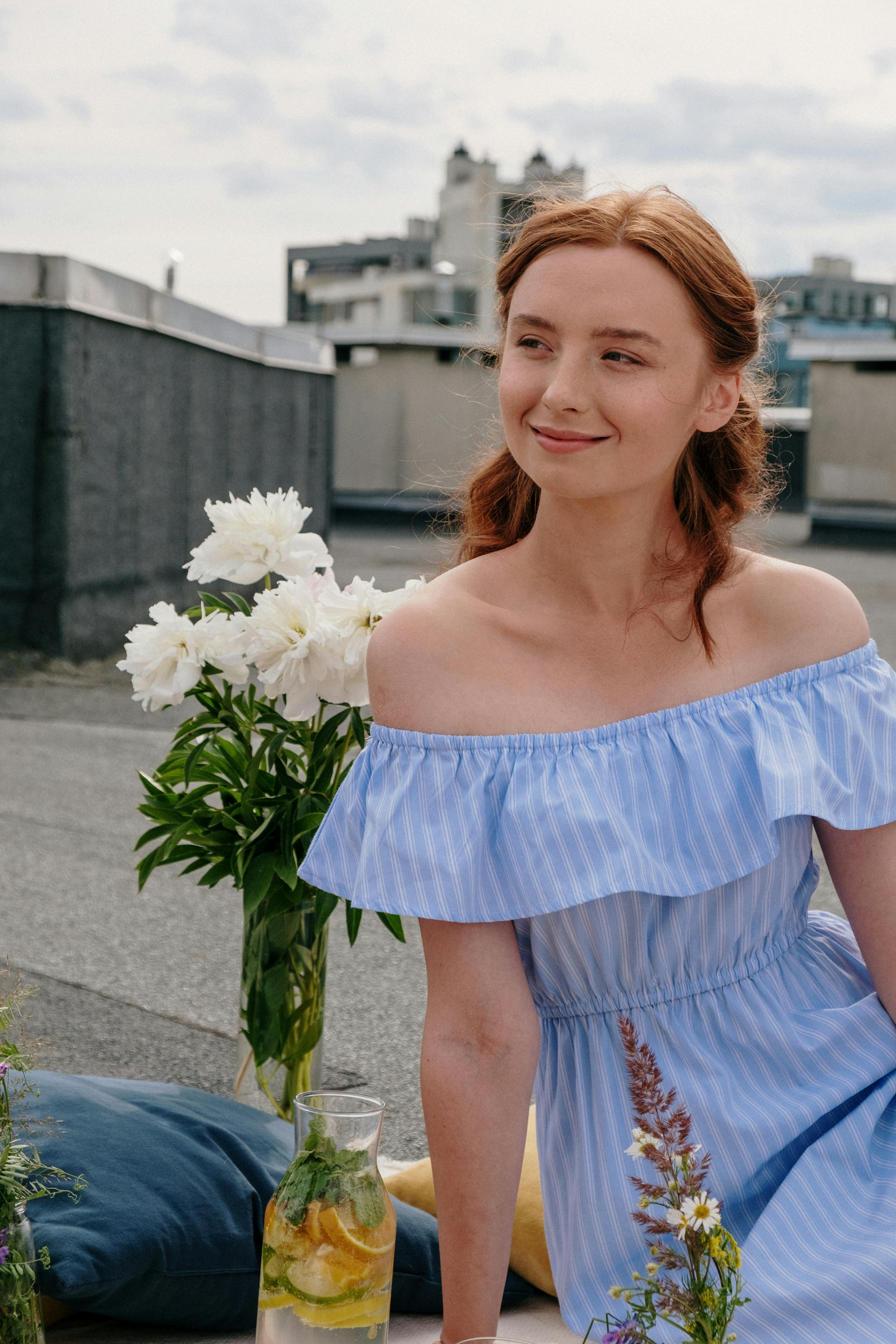 Portrait of a smiling redhead woman in a blue dress on a rooftop with flowers and lemonade, embracing summer vibes.
