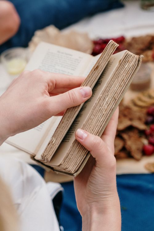 Free Close-up of hands holding and flipping through an aged book outdoors, surrounded by snacks. Stock Photo