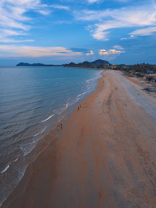 From above of distant unrecognizable travelers relaxing on sandy beach near waving ocean against cloudy blue sky