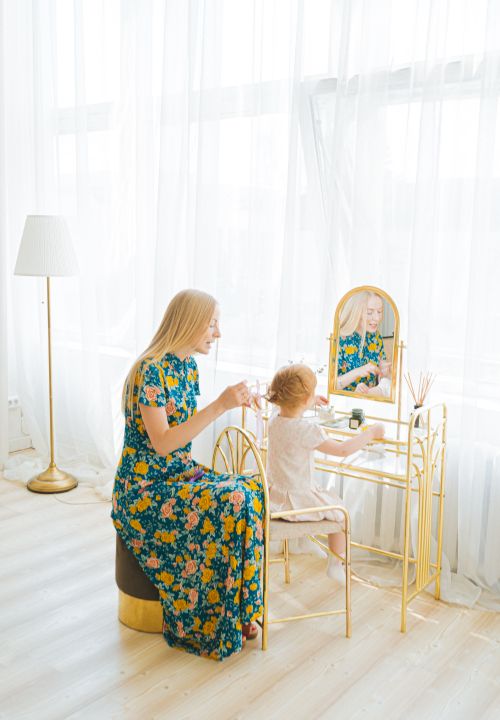 Free A mother in a floral dress braiding her young daughter's hair by a vanity table indoors. Stock Photo