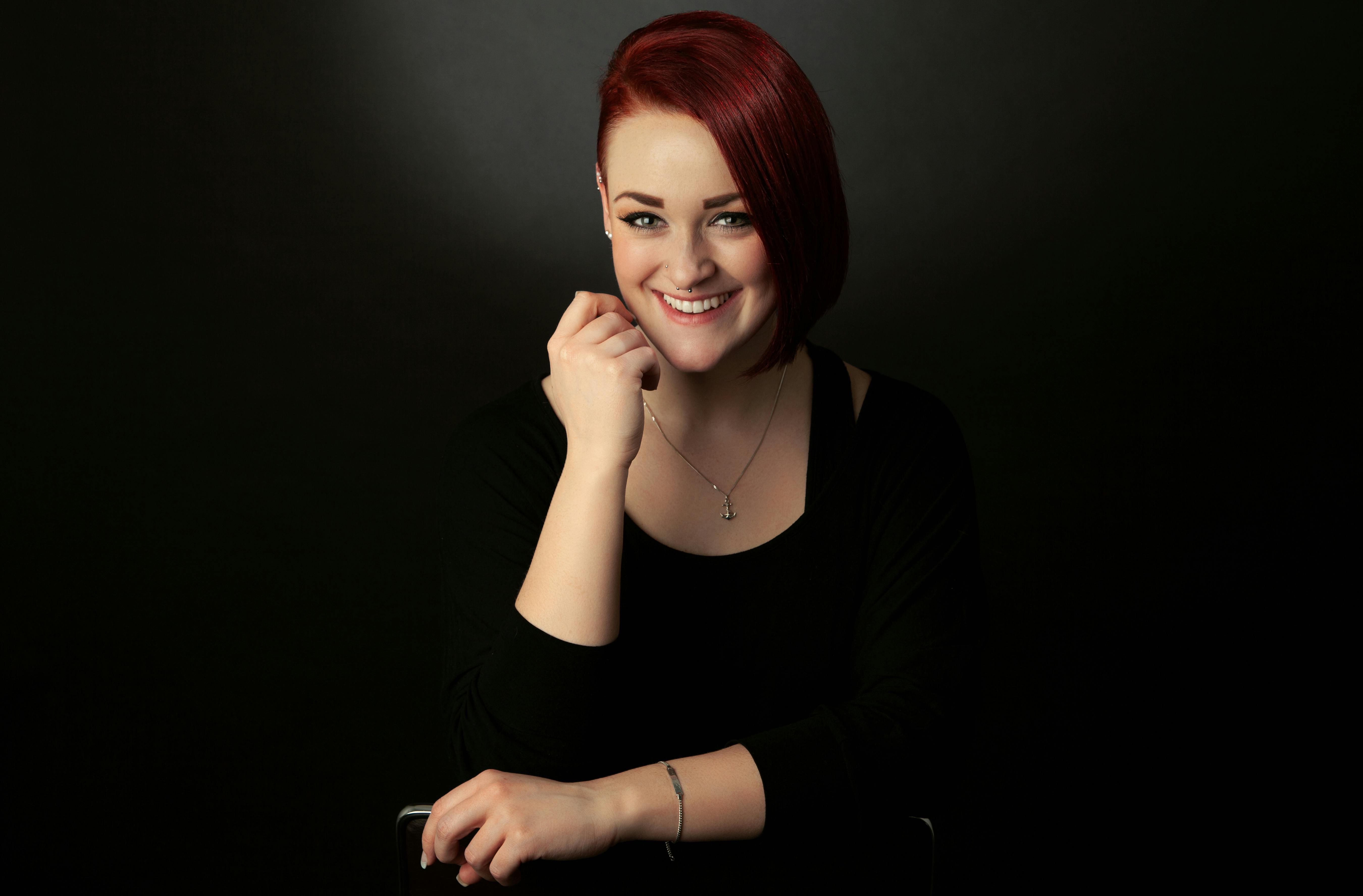 A smiling woman with red hair in a black outfit posing against a dark background in a studio.