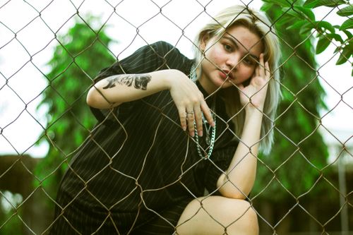 Free A young woman with a tattoo poses thoughtfully behind a chain link fence, surrounded by greenery. Stock Photo
