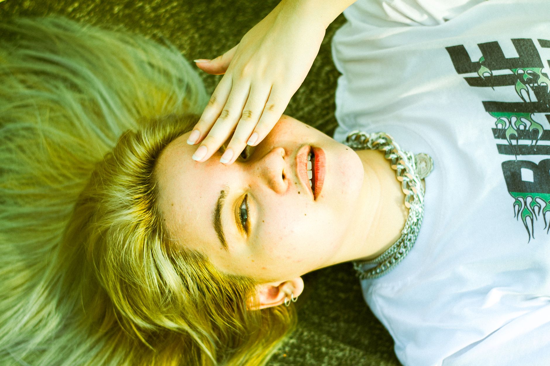 Young woman with blonde hair in a white shirt lying on grass covering her eye.