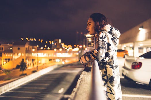 A young woman in a camo jacket leans over a railing, gazing at the illuminated cityscape at night.