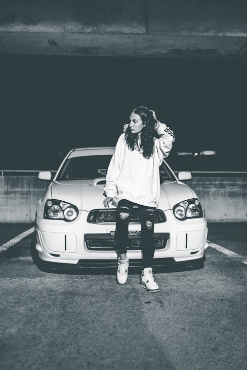 Black and white of attractive female with wavy hair and modern outfit sitting on car hood at night