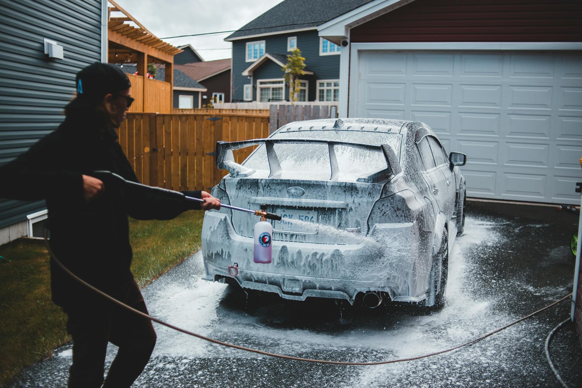 Side view of faceless male in black clothing washing car with foam in yard of house in daytime