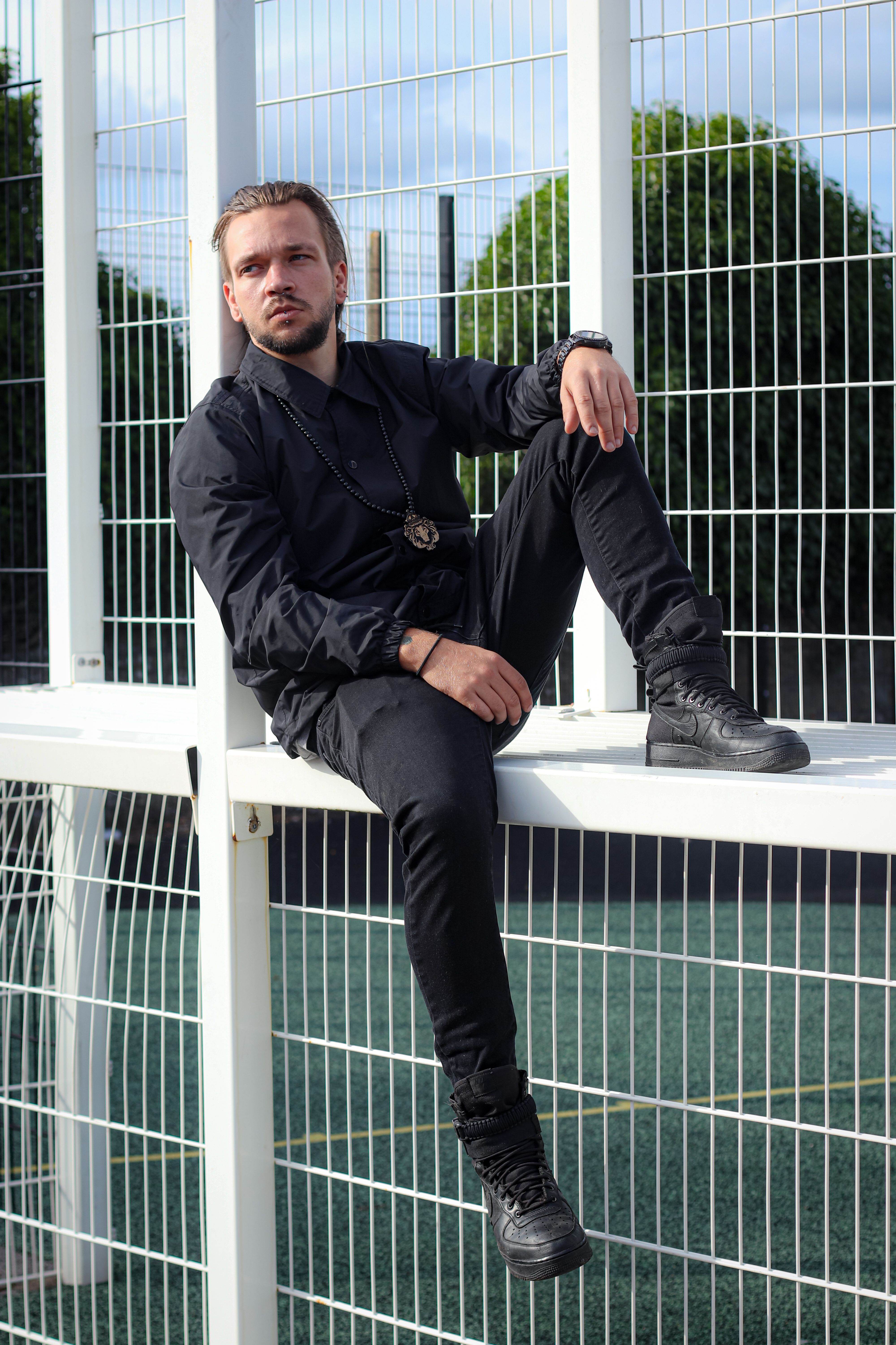 Full body of serious stylish male with brown hair and beard in black outfit sitting on white metal fence and looking away