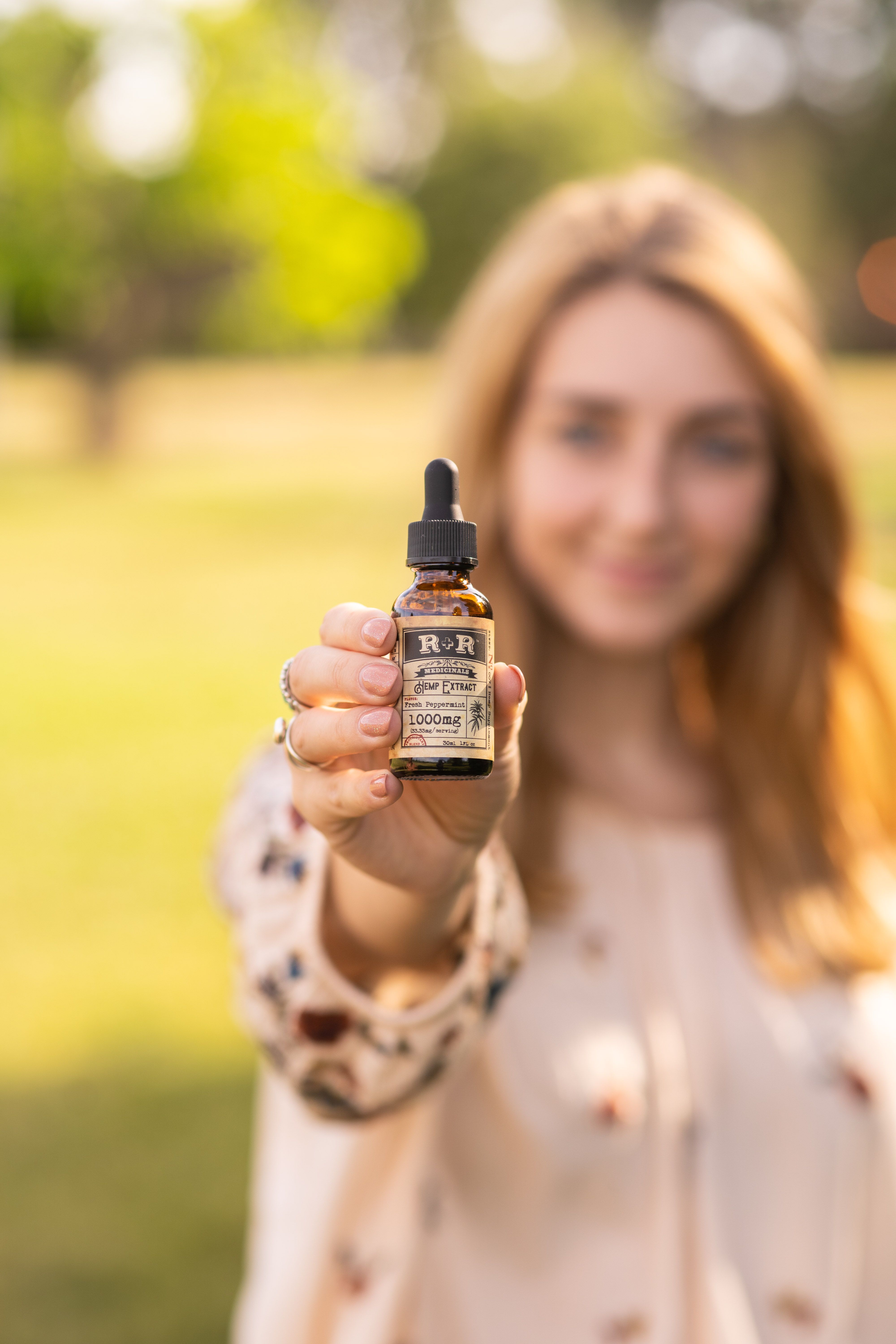 A woman holds an organic hemp serum bottle outdoors, showcasing natural skincare in Denver's park setting.