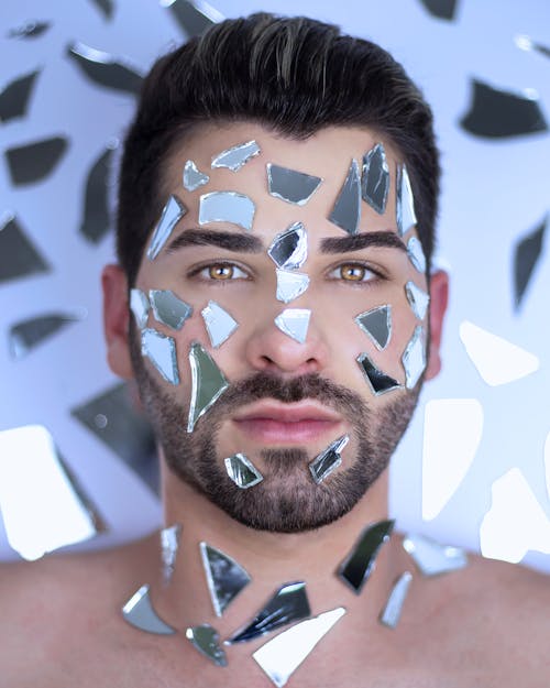 Calm man with dark hair and broken mirror pieces on face lying in surface with scattered chunks of glass while looking at camera