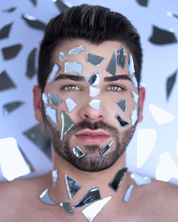 Calm man with dark hair and broken mirror pieces on face lying in surface with scattered chunks of glass while looking at camera