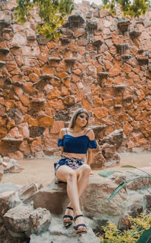 Woman in blue top and floral shorts posing against rustic stone backdrop.