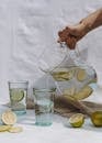 Crop person holding jug and pouring fresh lemonade into glass on white textile background