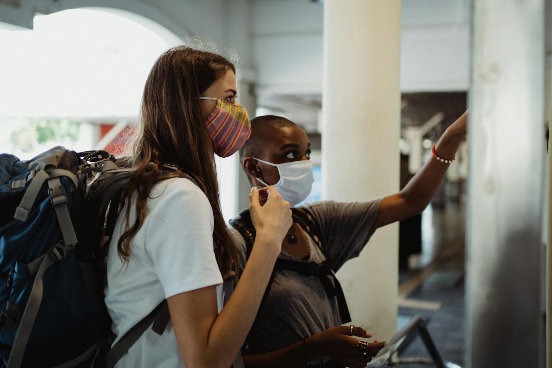 Two women in masks navigate a travel station with backpacks.