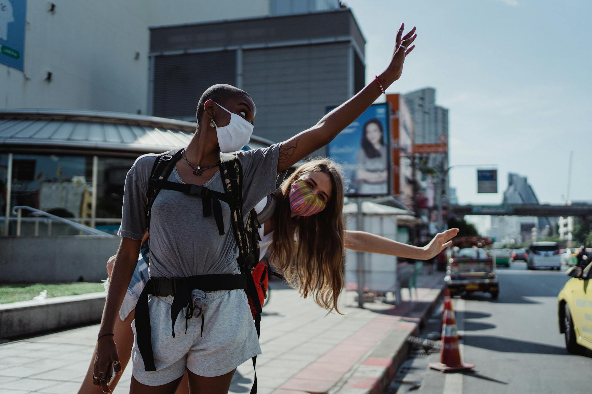 Two friends with backpacks waving in a bustling city, embracing travel adventure with safety masks.