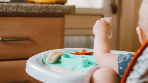 Baby in high chair eating food, surrounded by bananas in a kitchen setting.