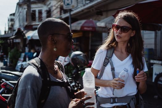 Two women with backpacks chatting and holding water bottles in a vibrant street market setting.