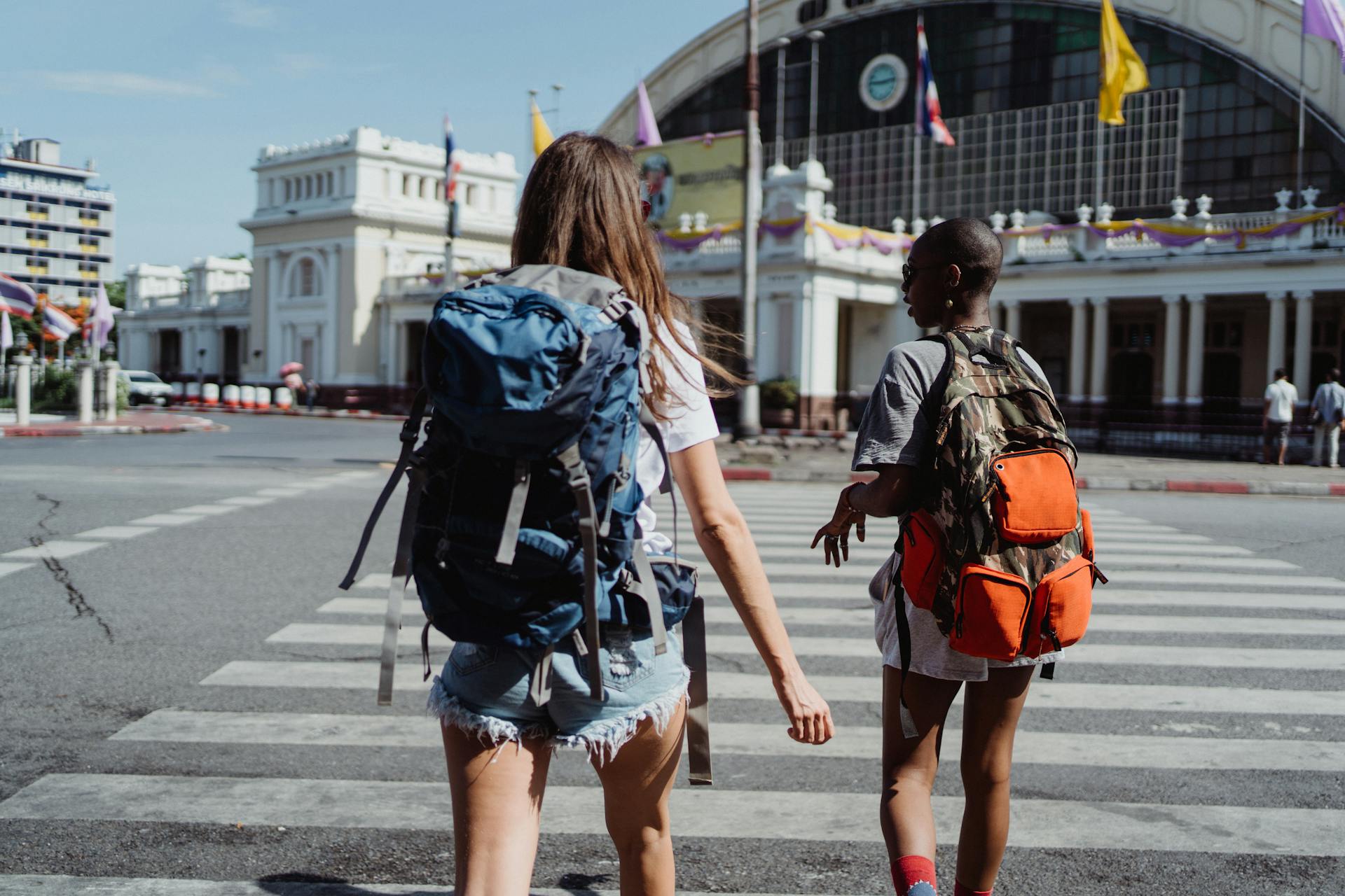 Two travelers with backpacks cross a city street near a historic building, symbolizing adventure and travel.