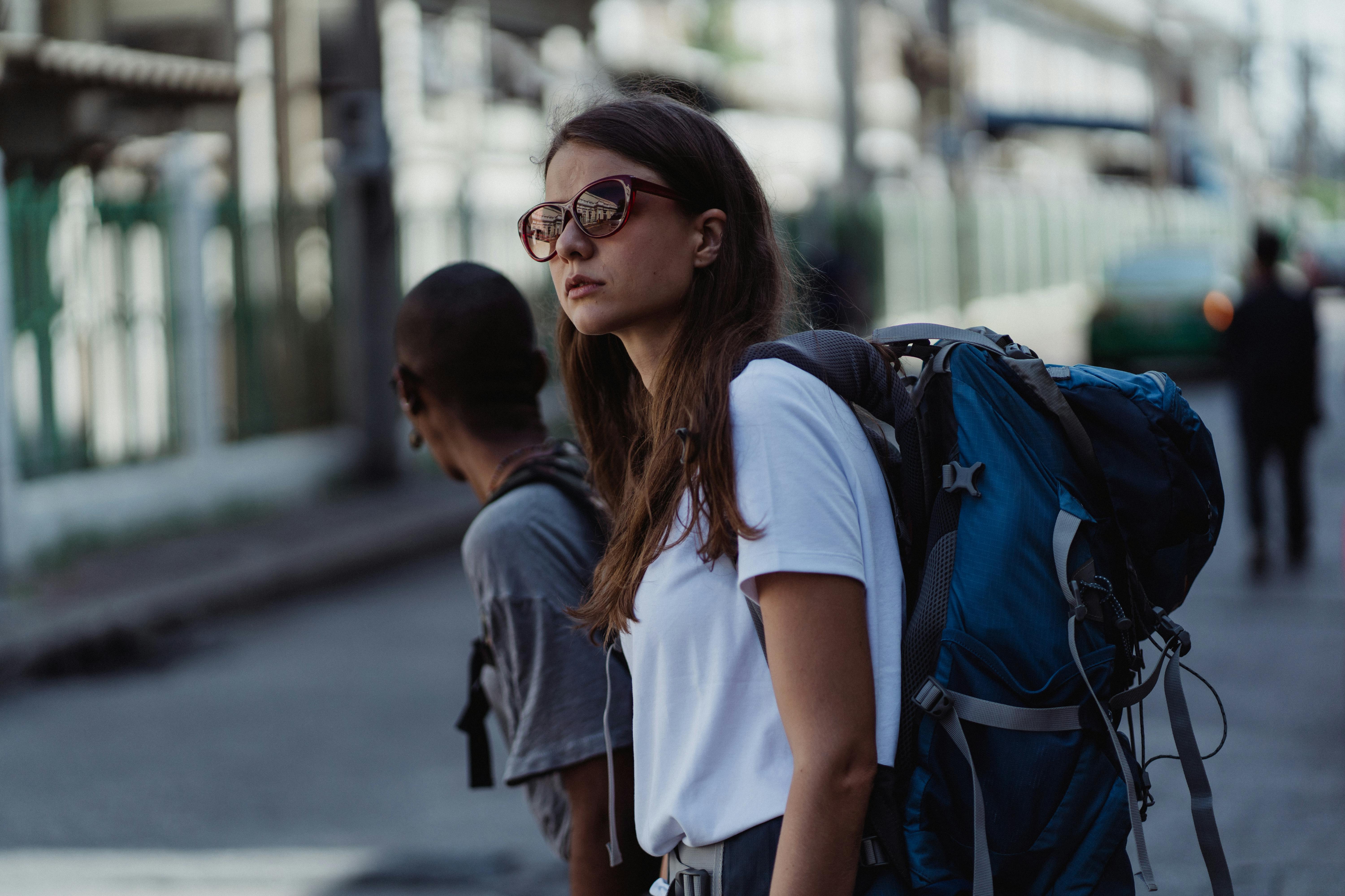 Woman wearing sunglasses and a large backpack standing on a city street with another traveler nearby.