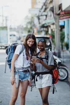 Two young women with backpacks using a cellphone while exploring city streets on a sunny day.