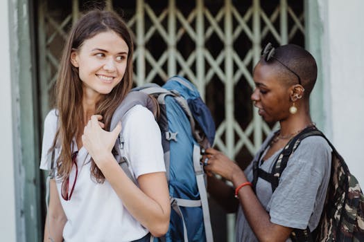 Two women preparing for travel with backpacks, smiling outdoors near a gate.