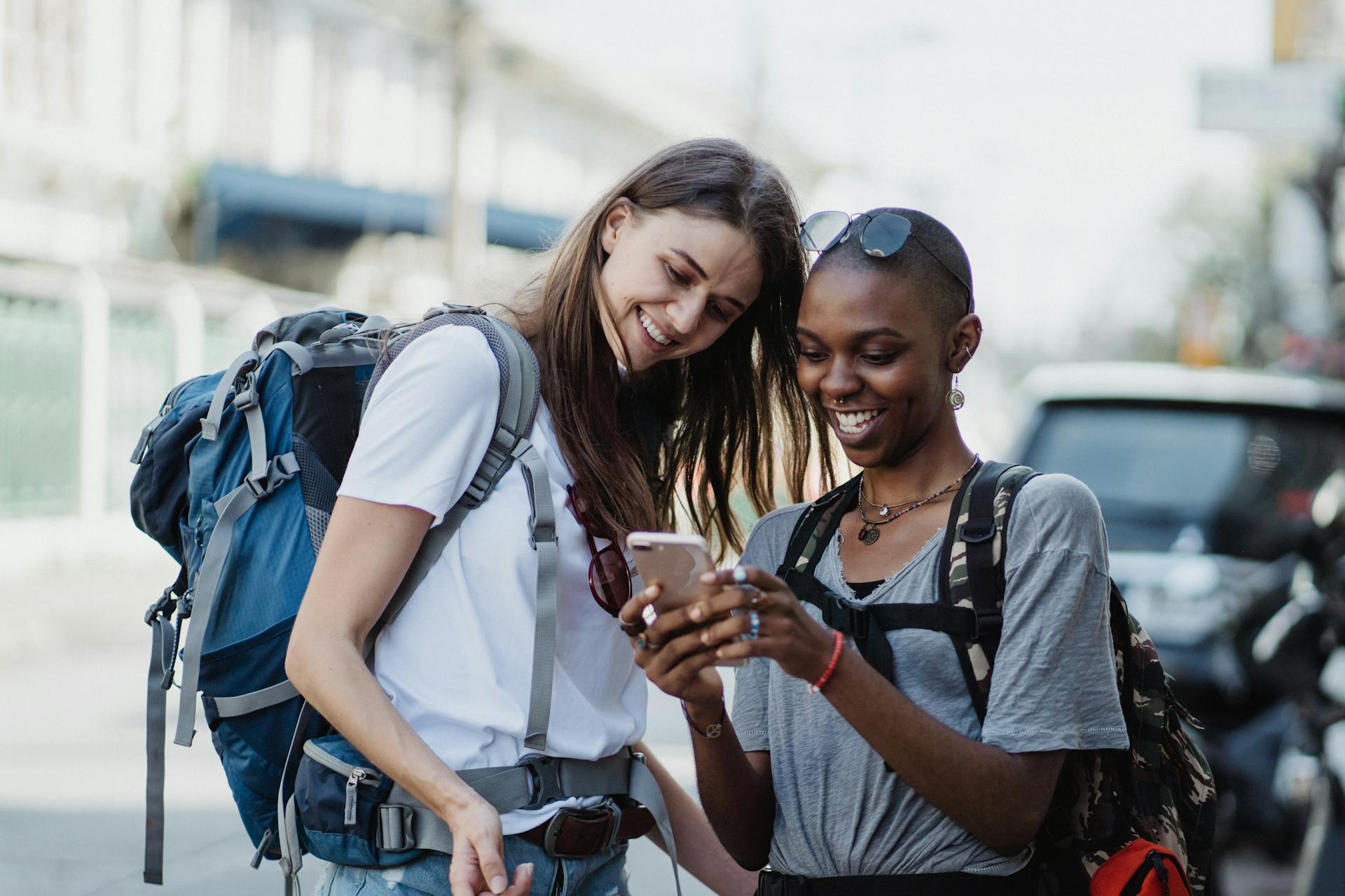 Two diverse women enjoying travel with backpacks, sharing a happy moment on a street.