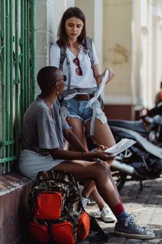 Two young women backpackers resting on a city street, examining a map, exploring an urban setting.