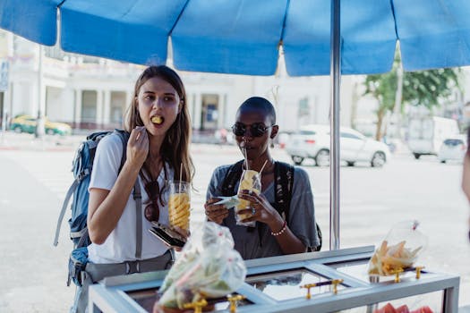Two women savoring fresh fruits at a street food stand under a blue canopy.