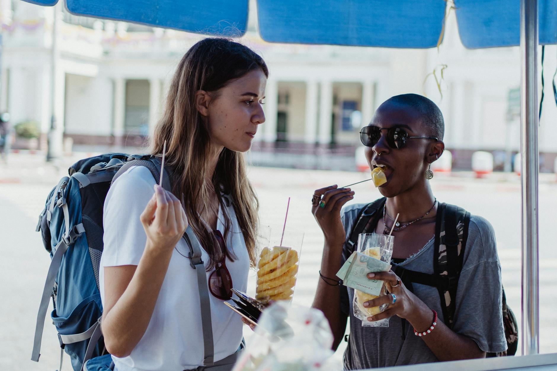 Two travelers eating fresh pineapple from street vendors in a lively urban setting