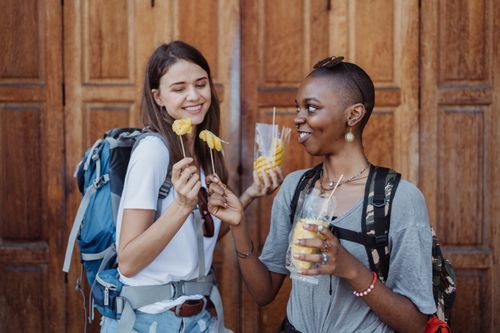 Free Two young women enjoying street food with backpacks, exploring a city street. Stock Photo