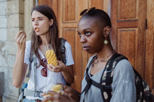 Two women savor fresh-cut pineapple while exploring a vibrant city street.