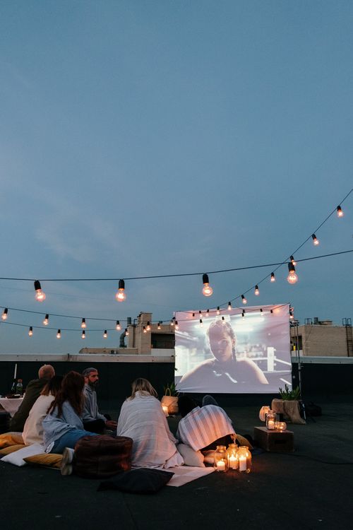 Free Group of friends enjoying a cozy rooftop movie night with string lights. Stock Photo