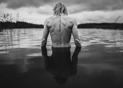 Black and white back view of shirtless man with tattoo standing in rippling lake surrounded by plants