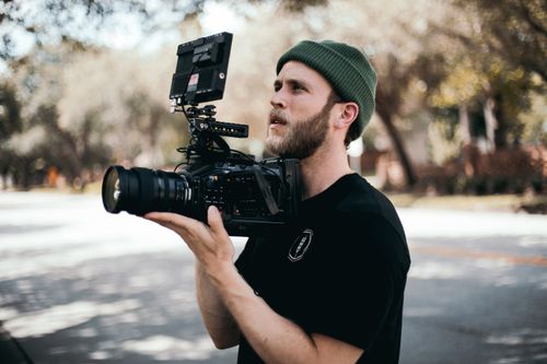 Free A focused bearded man in a beanie filming outdoors using advanced camera gear. Stock Photo