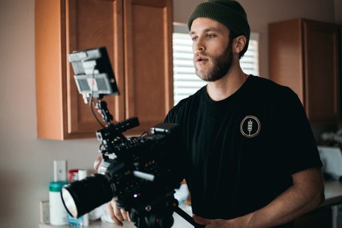 Free A bearded man in a beanie operates a professional video camera inside a room. Stock Photo