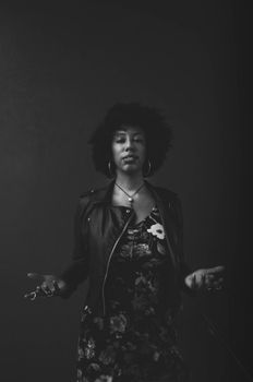 black and white of serious confused young African American lady in leather jacket standing against dark background and looking at camera