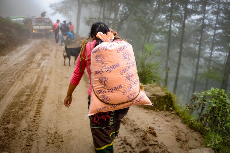 Back View Of A Person Carrying A Sack On His Back While Walking On The Muddy Ground