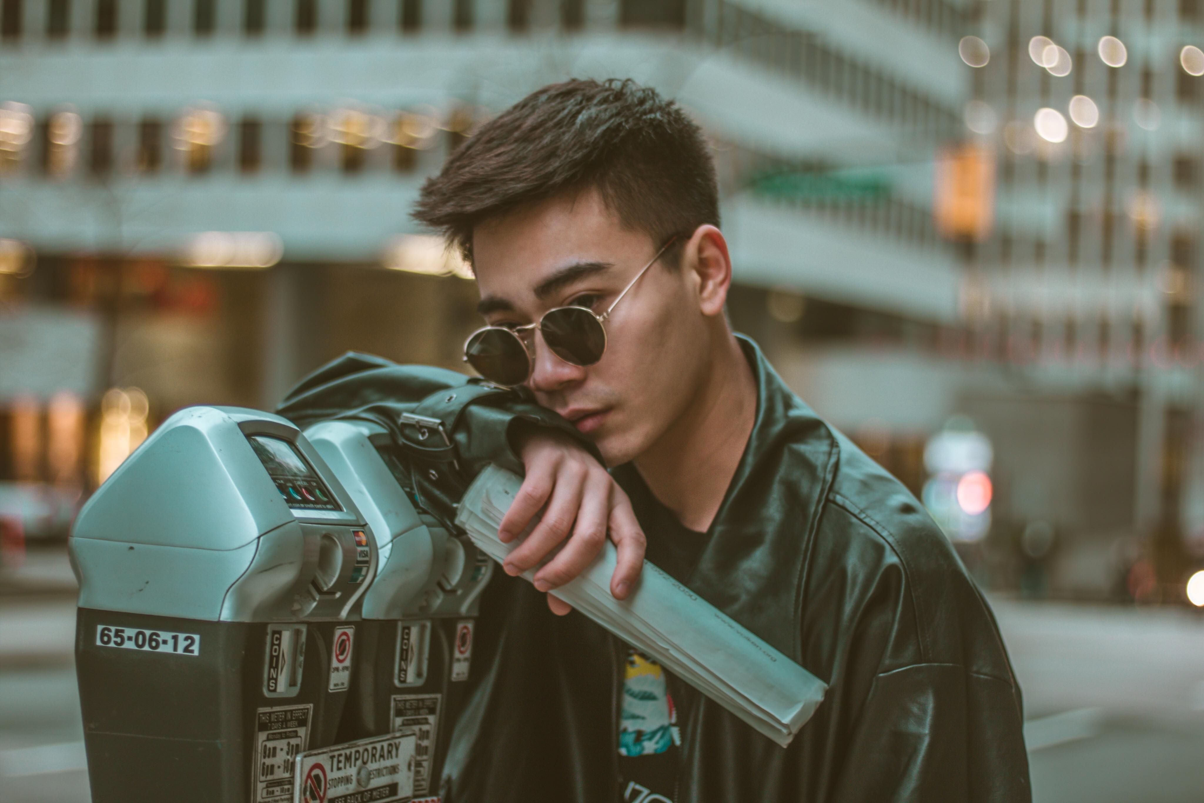 Fashionable young man in sunglasses and leather jacket posing against cityscape.