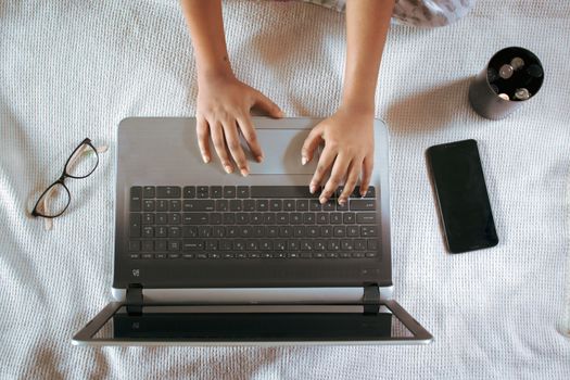 From above of crop unrecognizable female remote employee working on netbook near cellphone and eyeglasses on crumpled bed sheet