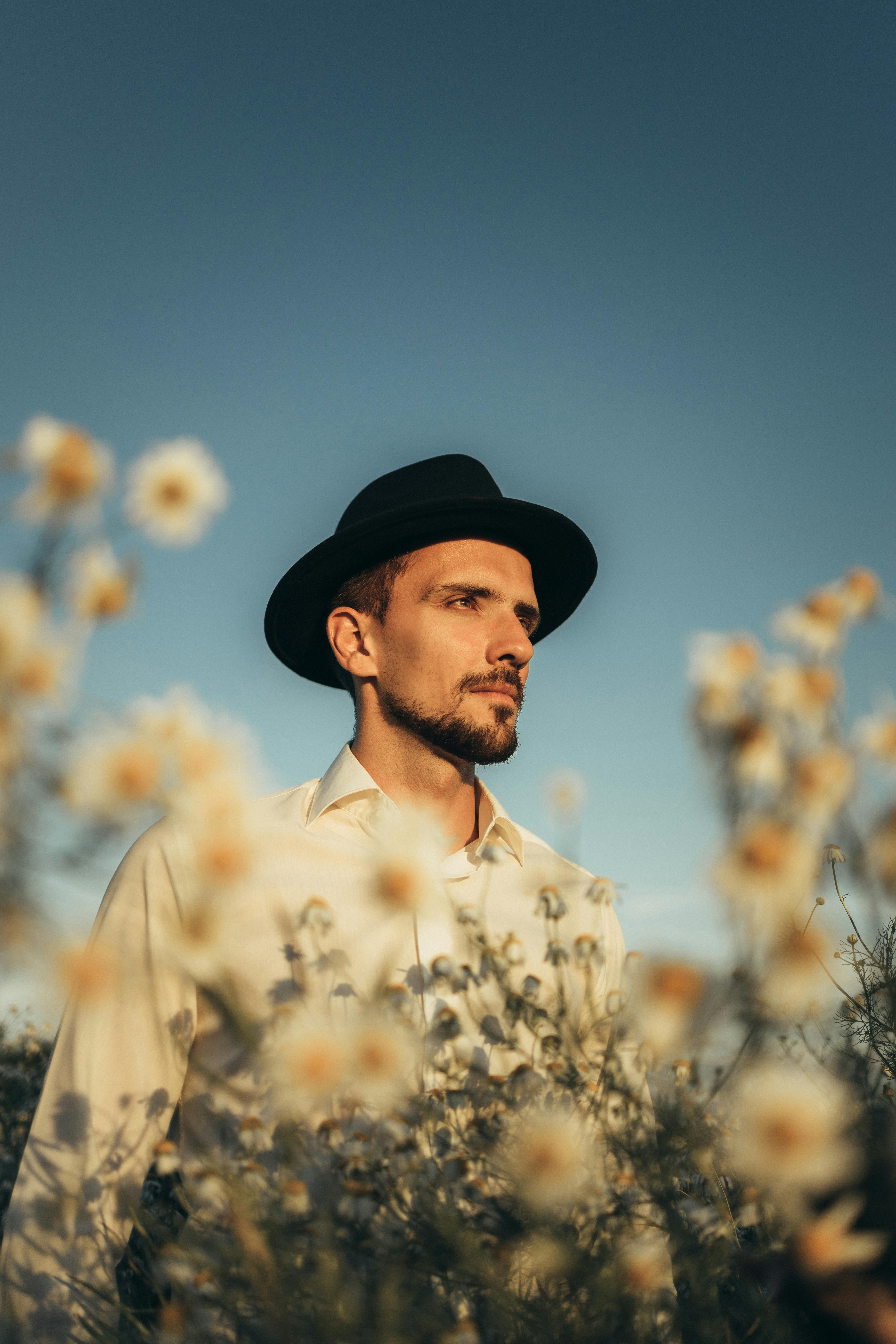 一位时尚男子头戴软毡帽，站在野花田中，身后是宁静的蓝天