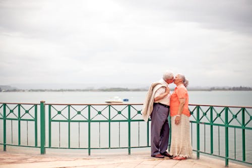 Side view full body romantic senior couple wearing trendy outfits standing on waterfront promenade and kissing gently on overcast weather