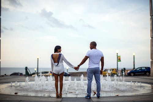 Back view full body positive African American couple in stylish summer wear holding hands and looking at each other while standing near fountain on seafront