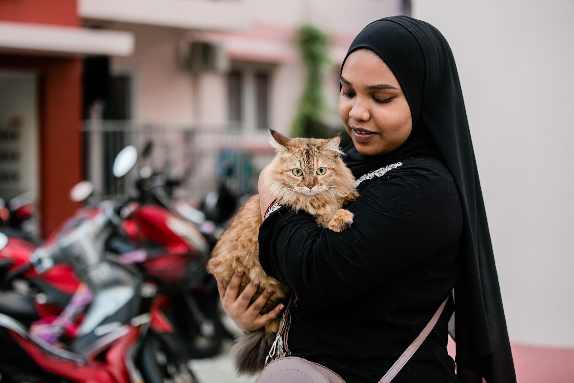 A smiling woman in a hijab holds a tabby cat outdoors, surrounded by motorcycles.