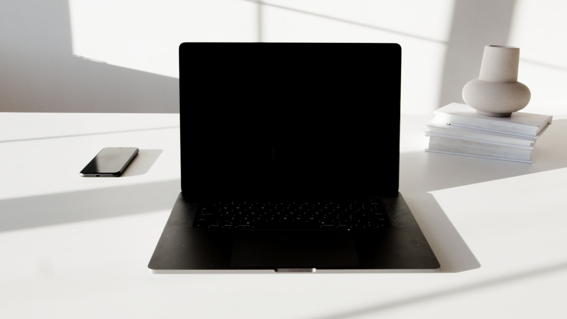 A minimalist white desk featuring a laptop, smartphone, and decorative vase in a modern setting.