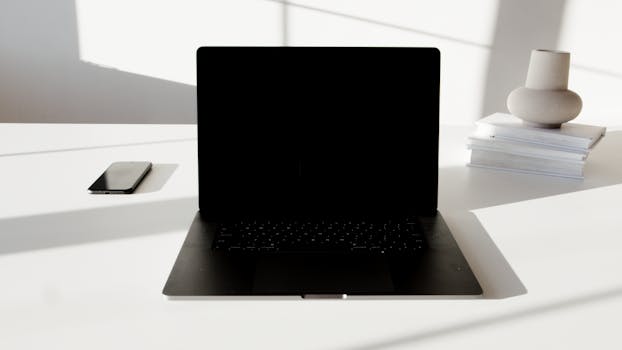 A minimalist white desk featuring a laptop, smartphone, and decorative vase in a modern setting.