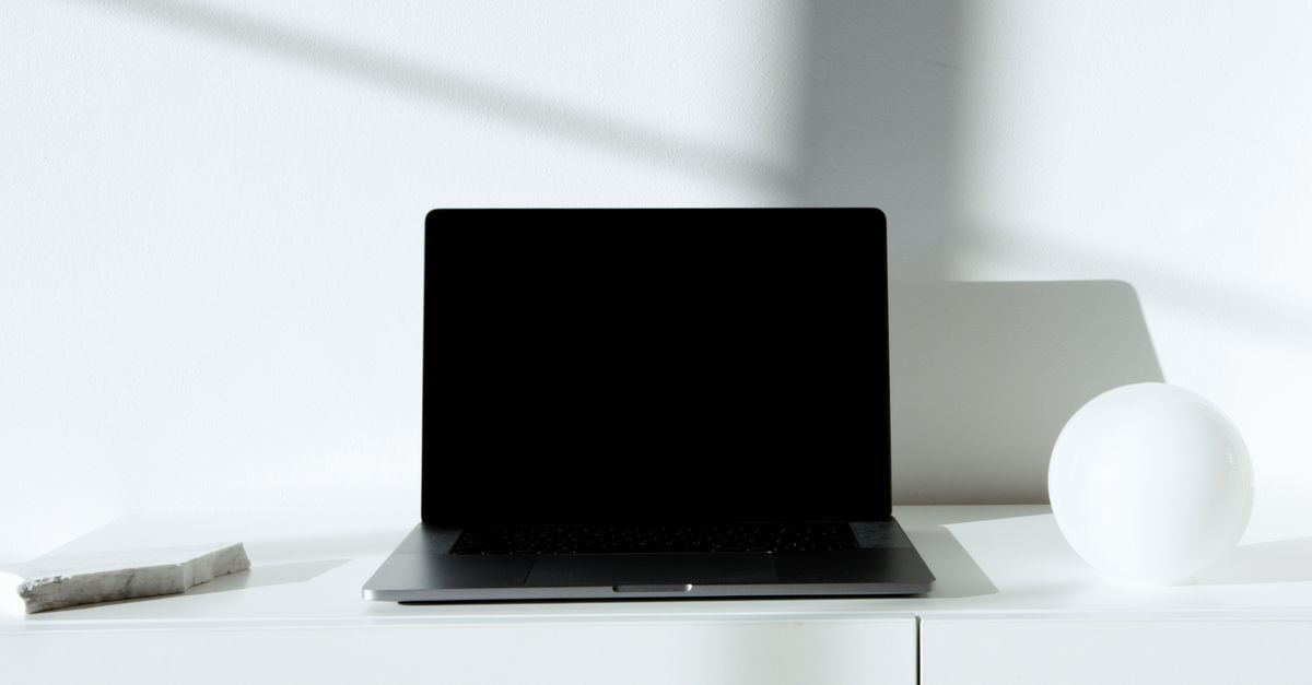 A sleek black laptop on a white desk in a minimalist setup with natural lighting.