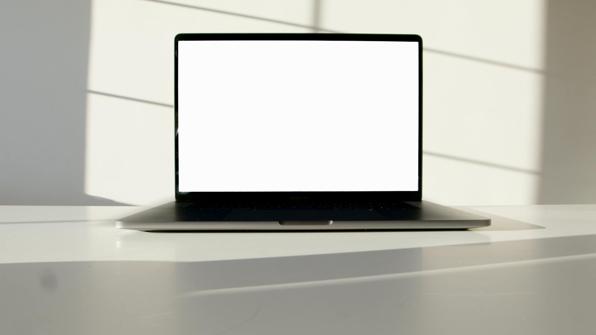 Laptop with blank screen on a white table in sunlit room, perfect for mockup presentations.