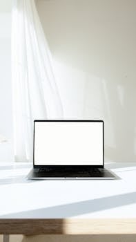 A minimalist laptop with a blank screen sitting on a bright white desk in a sunlit room.