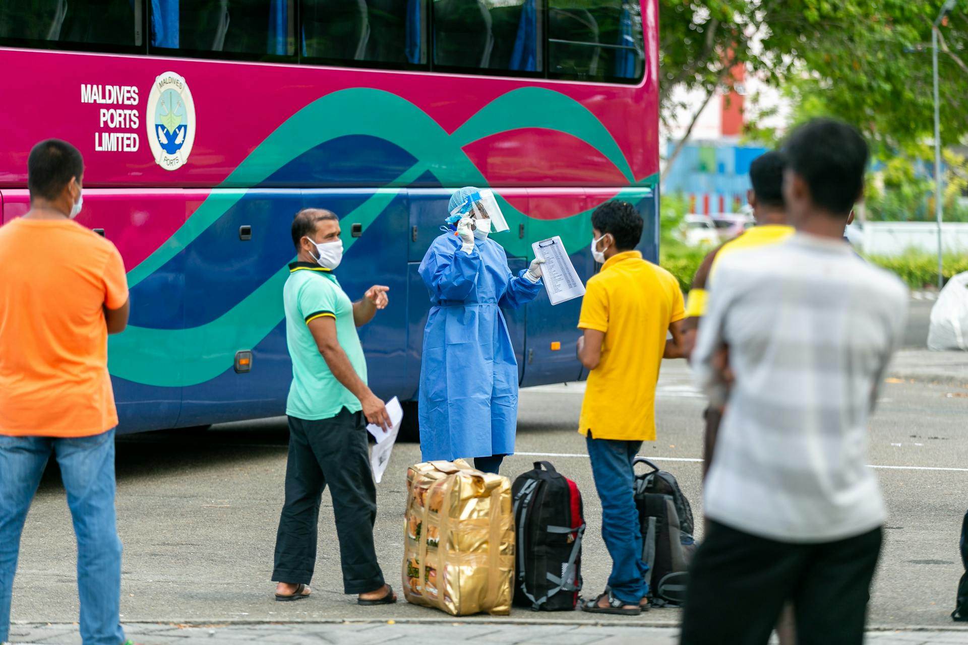 A healthcare worker in PPE assists passengers near a colorful bus, emphasizing safety protocols.