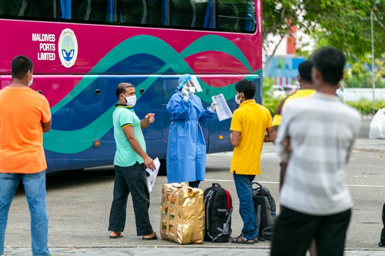 A Person In PPE Standing With Passengers By The Side Of A Bus