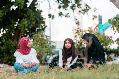 Full body cheerful young Muslim females wearing traditional hijab sitting on grassy meadow and having conversation in park